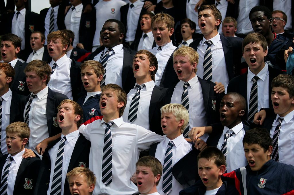 A diverse group of schoolboys in uniforms cheering energetically outdoors.