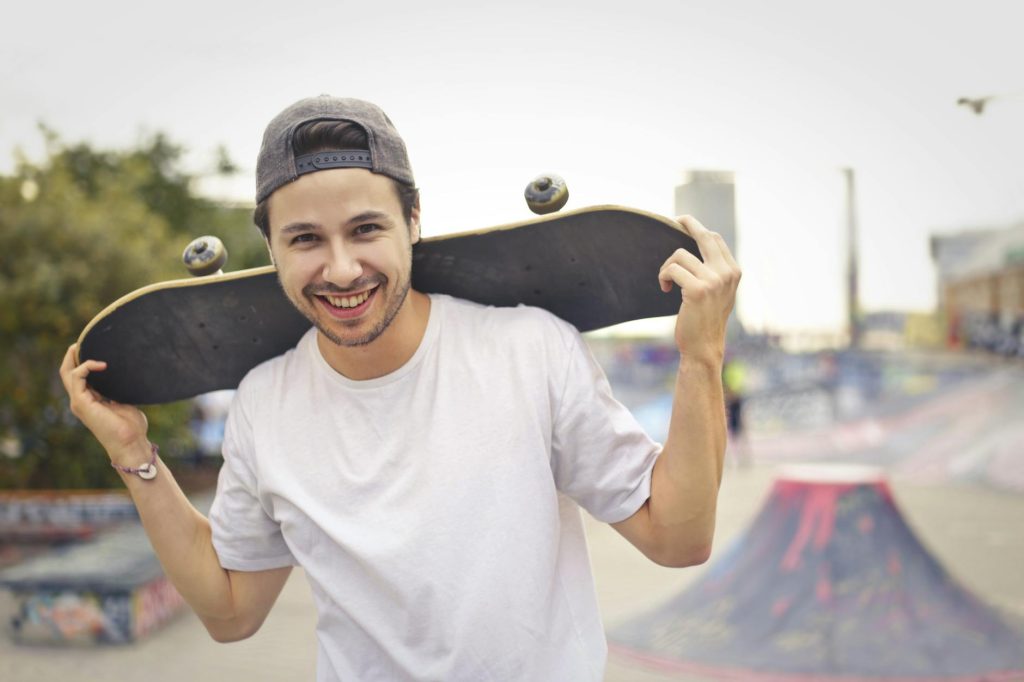 Happy skater with a skateboard over shoulders, smiling in a park setting.