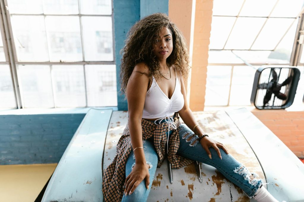 Young woman sitting on a vintage rusty car hood in a stylish loft with large windows.