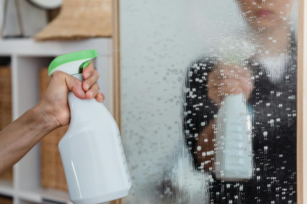 Close-up of a hand using a spray bottle to clean a mirror indoors.