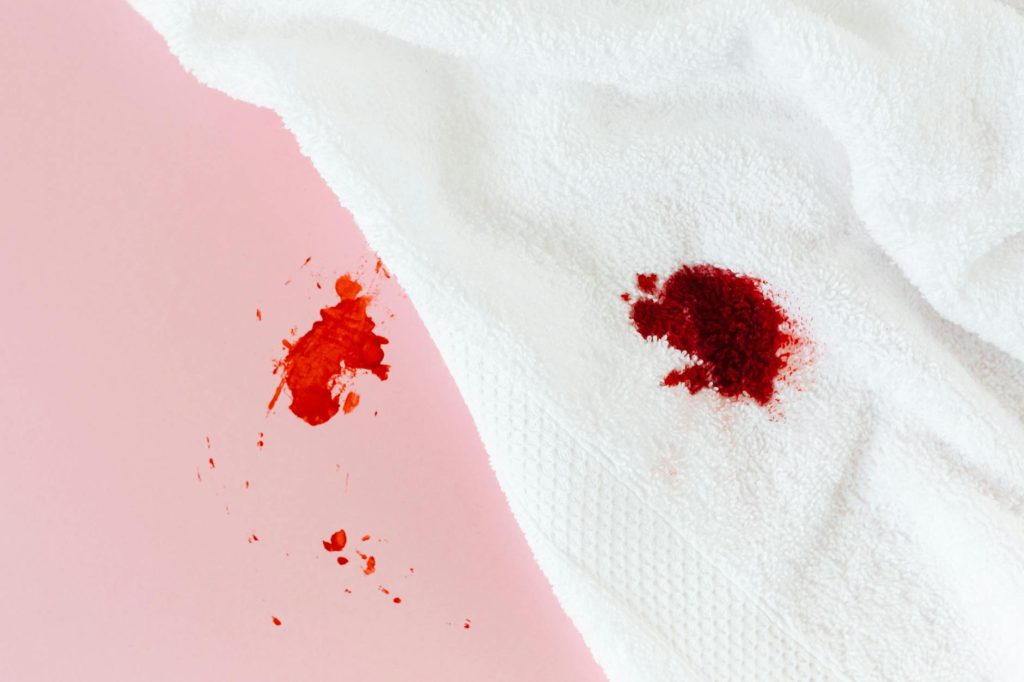Red blood stain on a white towel spread over a pastel pink background, close-up shot.