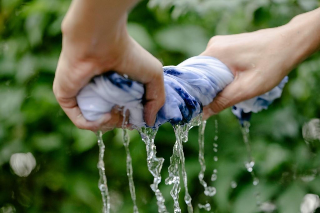 Close-up of hands squeezing blue tie-dyed fabric, water flowing outdoors.