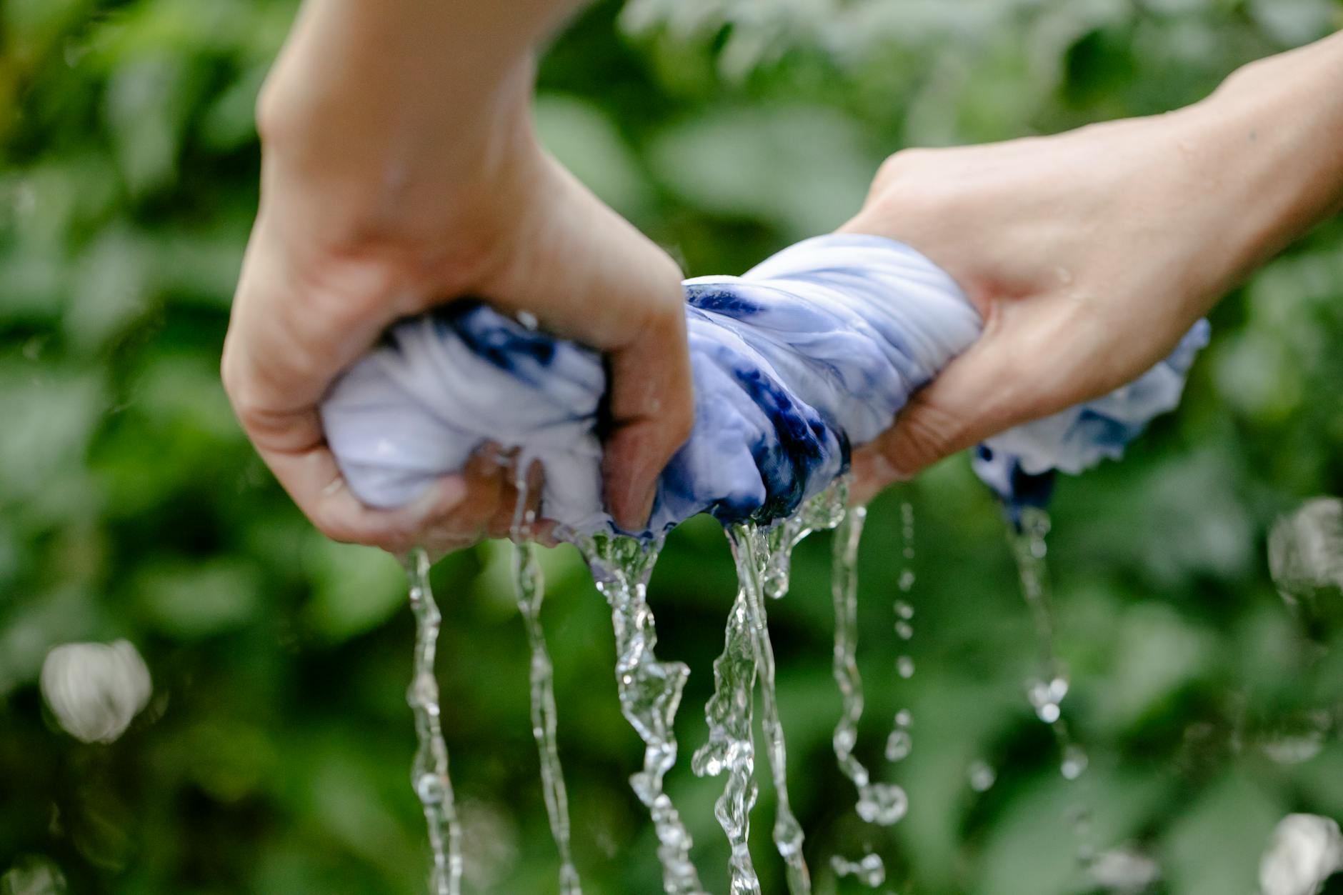 Close-up of hands squeezing blue tie-dyed fabric, water flowing outdoors.