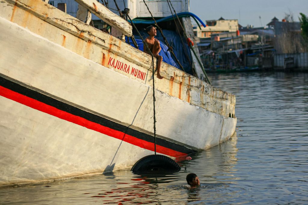 A young boy perched on a ship at Sunda Kelapa harbor in Jakarta, Indonesia. Serene waters and local life.