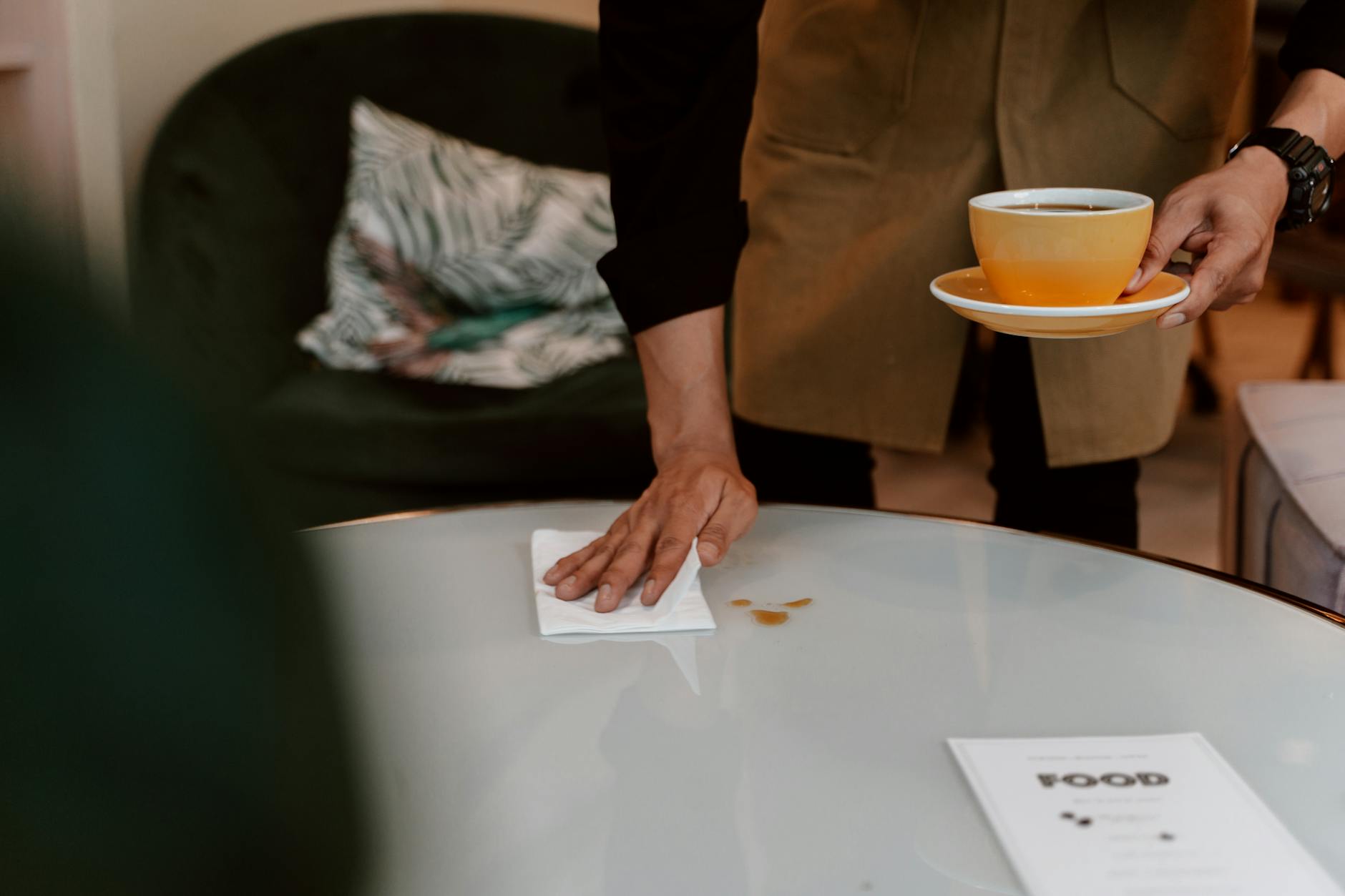 A barista wiping a table clean while holding a ceramic cup in a cozy cafe setting.