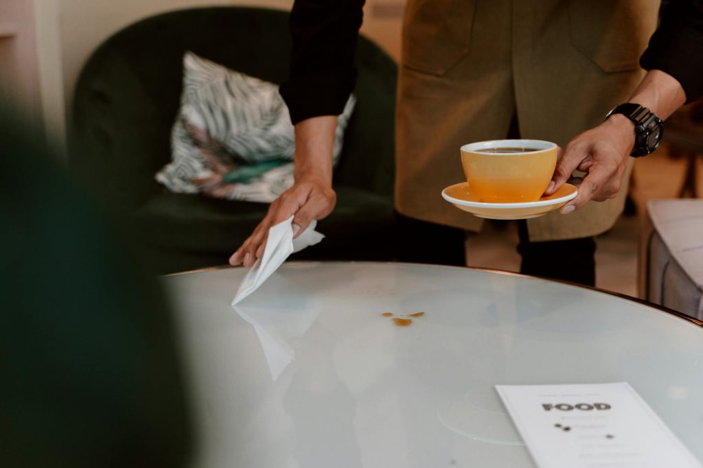 A barista holding a coffee cup and cleaning a table in a cozy cafe setting.