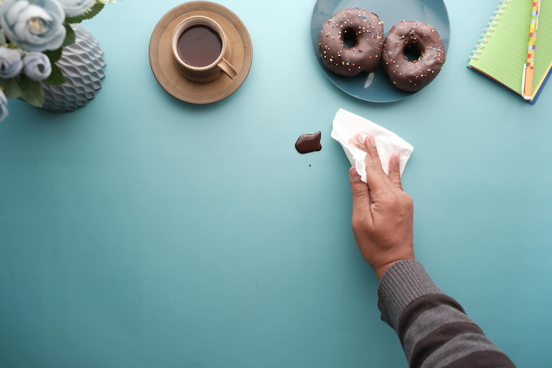 A hand wipes chocolate from a blue table beside coffee and donuts.