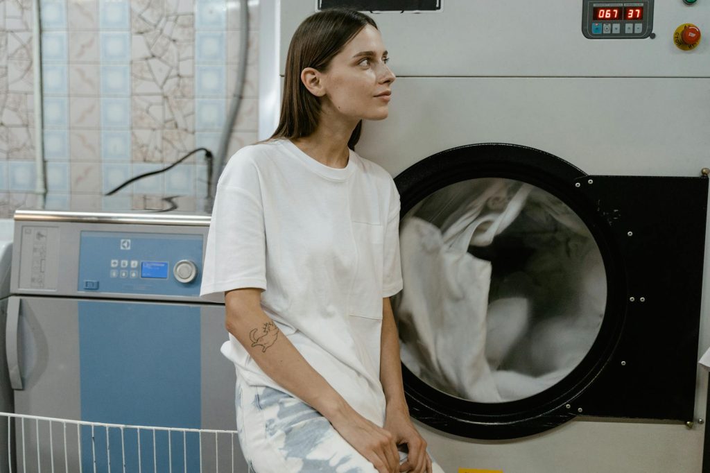 A young woman in a white shirt leans against an industrial washing machine in a laundry facility.