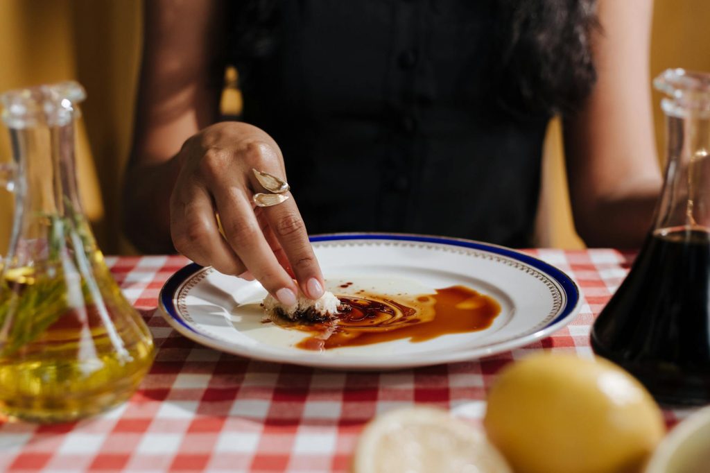 A woman dips bread in balsamic glaze on a checkered tablecloth, capturing a cozy Italian dining experience.