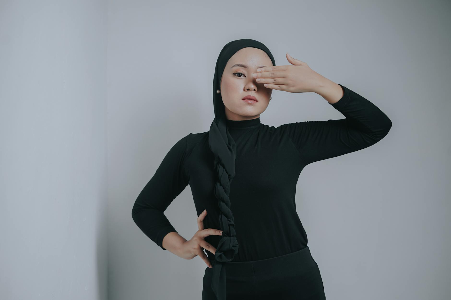 A fashionable woman poses in studio with hijab, covering one eye, in a modern minimalist style.