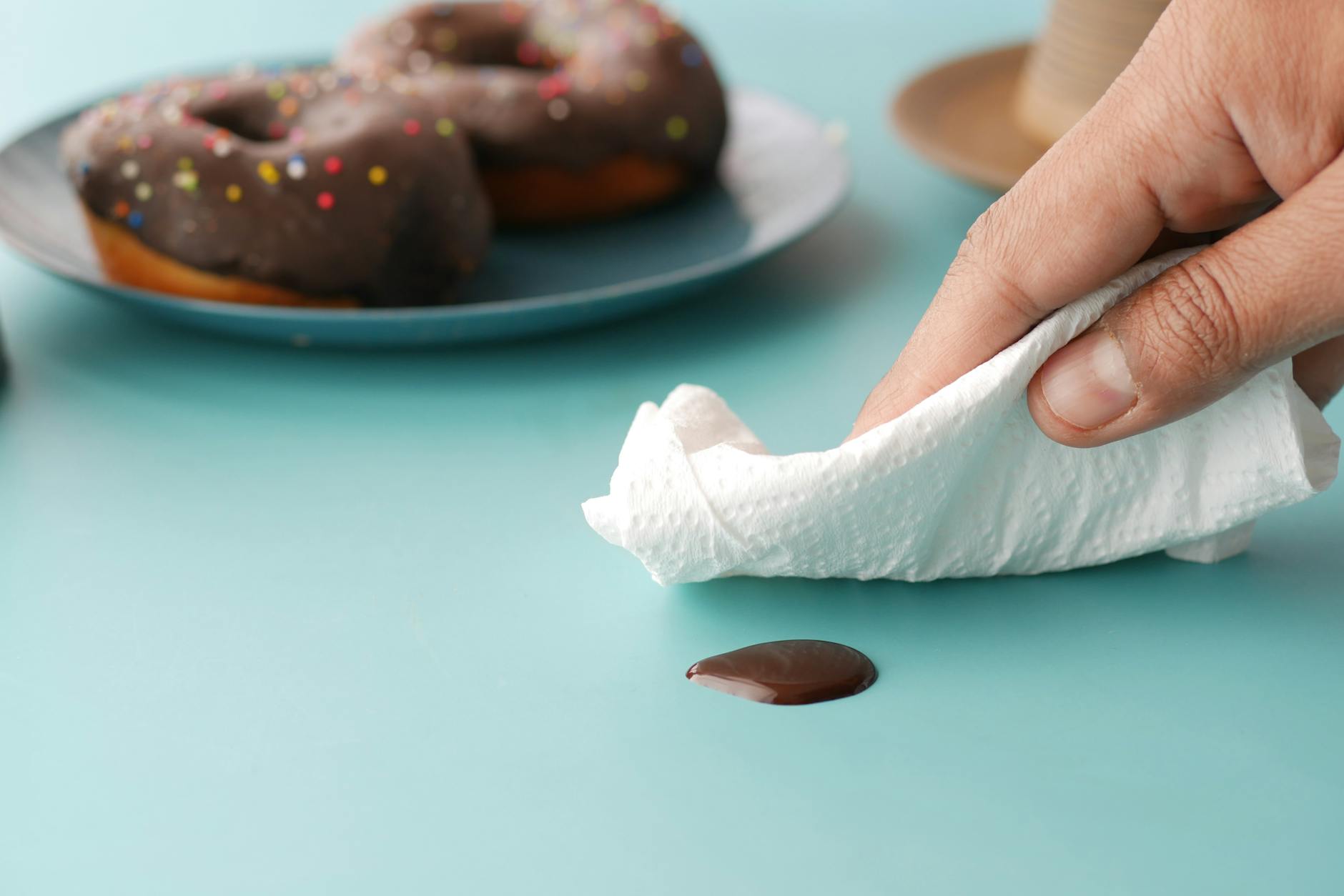 Hand wiping chocolate spill with tissue near doughnuts on a blue table.