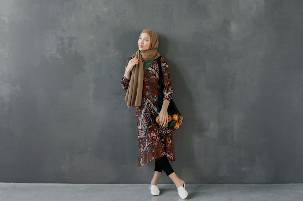 Stylish woman in a hijab posing against a textured wall, showcasing traditional wear.